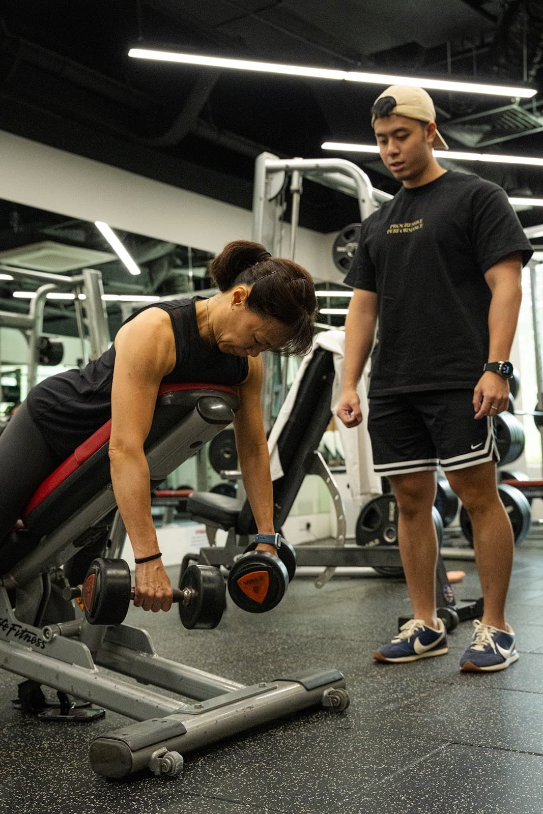 A woman supported on an incline bench holding two dumbbells while a Progressive Performance coach observes.