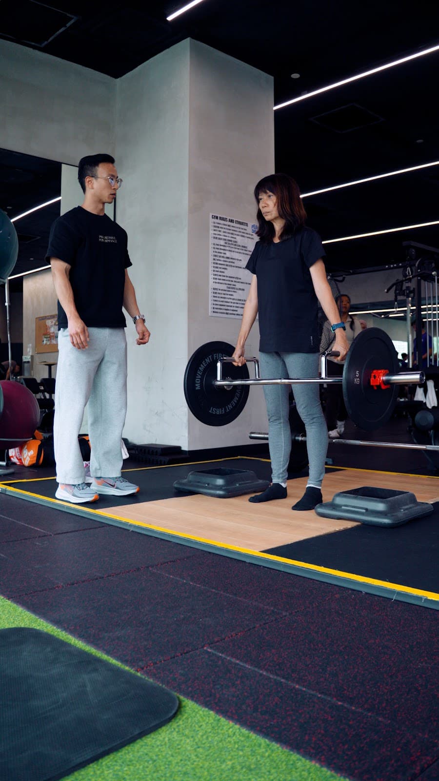 A woman holding a loaded trap bar with a Progressive Performance coach standing beside her.